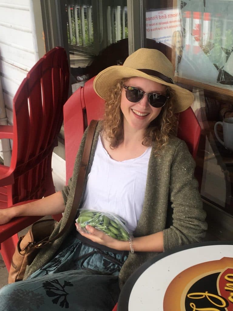 Young woman holding a bag of string beans, sitting down in a red chair, wearing a summer hat and sunglasses.