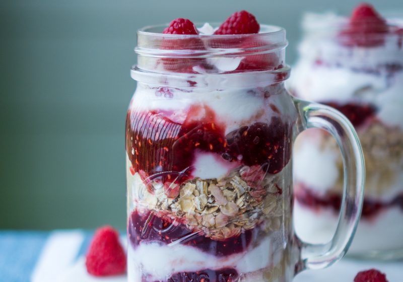 two mason jars layered with white yoghurt and red berries, on a blue background