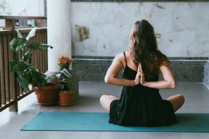 Woman showing her back with her arms bent behind her back for a yoga pose. She is sitting on her yoga mat on a balcony with flowers.