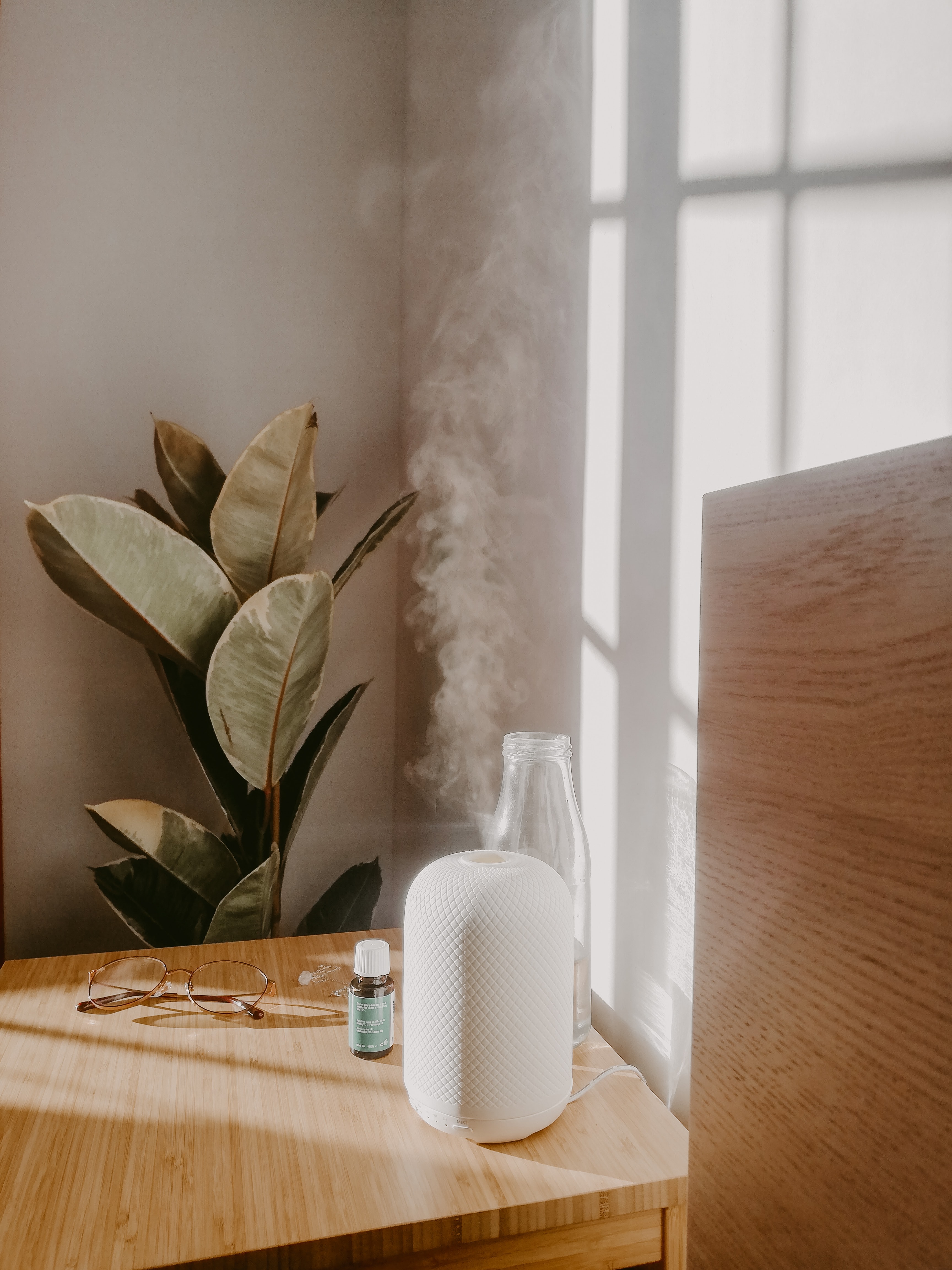 White essential oil diffuser on a wooden table. There are glasses and an essential oil bottle on the table, and a tropical plant is located behind the table.