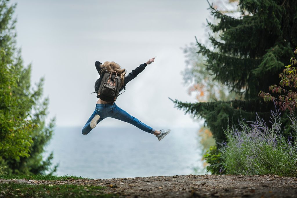 Girl with long hair wearing a backpack and blue jeans is jumping in the air, facing the ocean and surrounded by trees