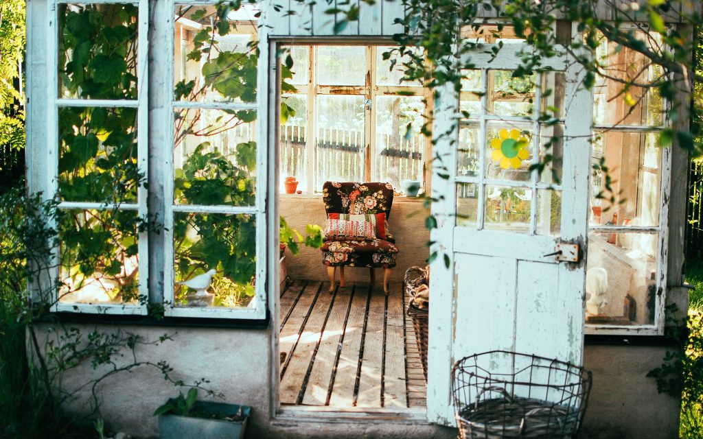 Old sunroom made of glass windows and wood with wood floorboards, and covered in plants.