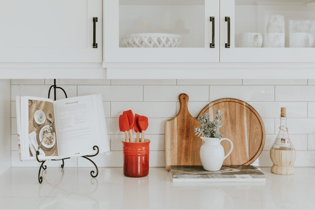 White kitchen counter with white tiles and white cupboards with white dishes. A cutting board, a cook book, red cooking utensils and a cookbook are displayed on the counter.