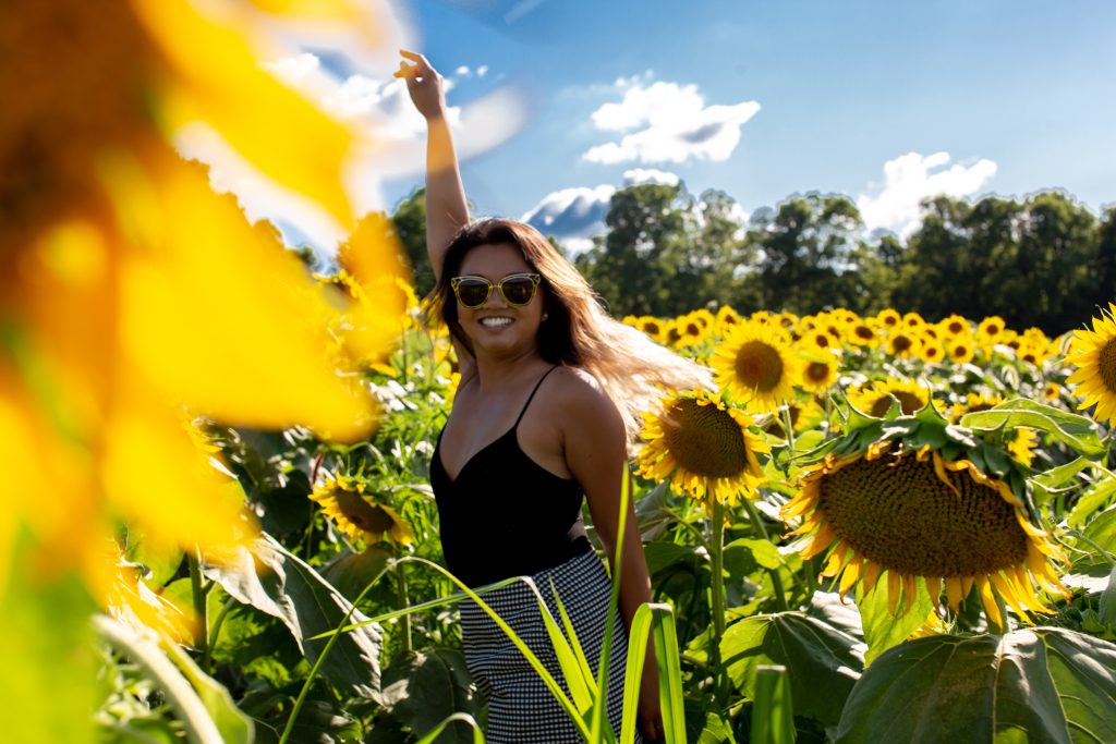 Girl with sunglasses and a black tank top smiling and waving in a field of sunflowers.