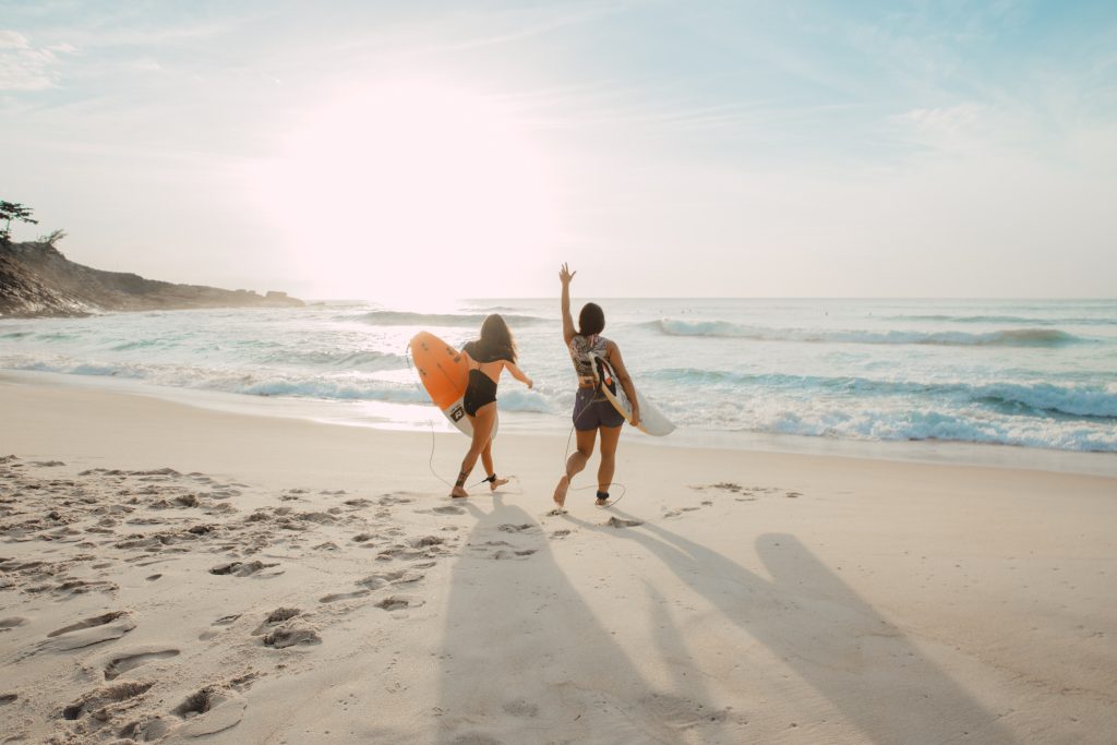 Two girls carrying surfboards waves to the sun as they walk on the beach towards the ocean waves.