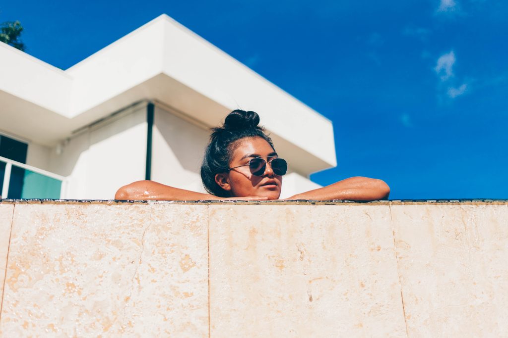 Girl with black hair in a top bun wearing sun glasses looks over a cement wall, with a blue sky and white house in the background.