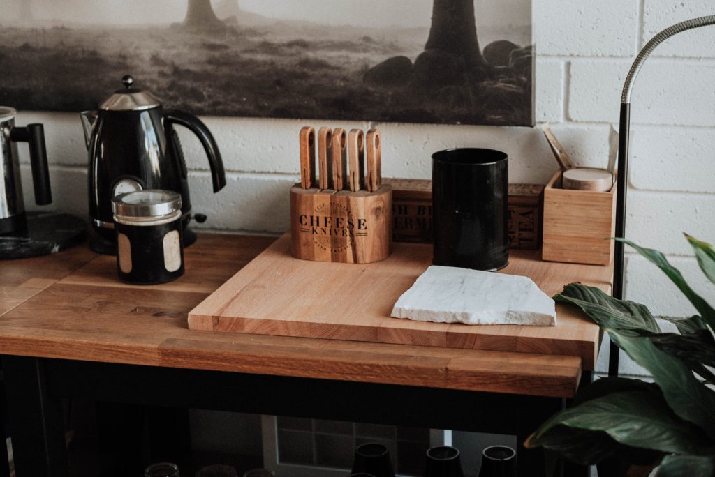 Cutting board counter with more cutting boards, a copper knife set, a kettle, and a plant and artwork in the background. Cups and glasses are hidden beneath the wooden counter.