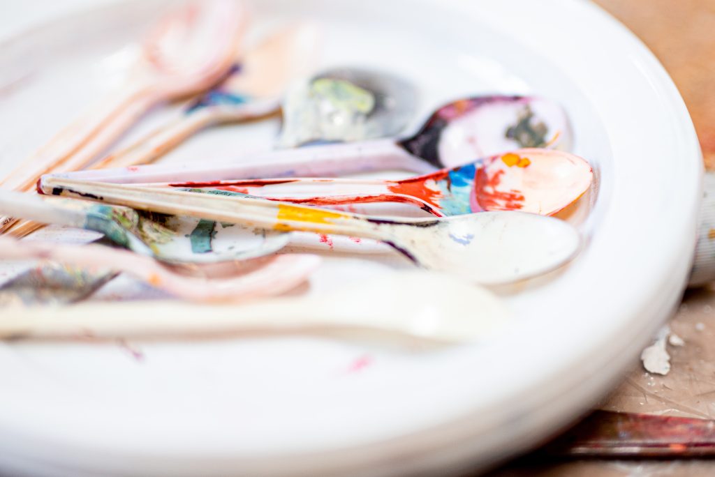 Many plastic spoons splattered in different colours of pain on a white plate.