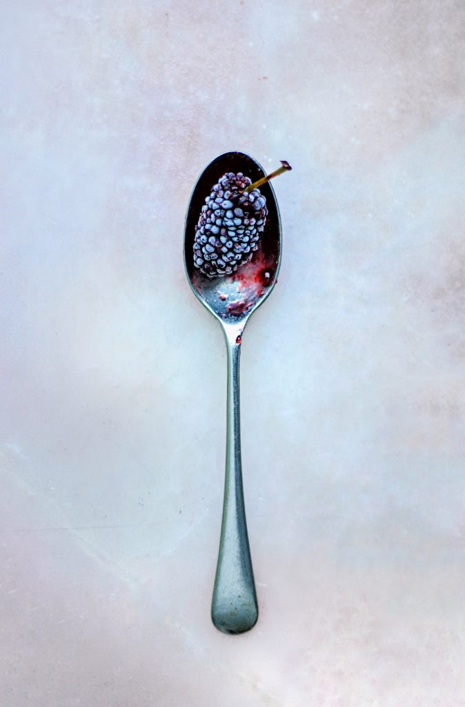 Silver spoon holding a mulberry on a white and pink marbled background.