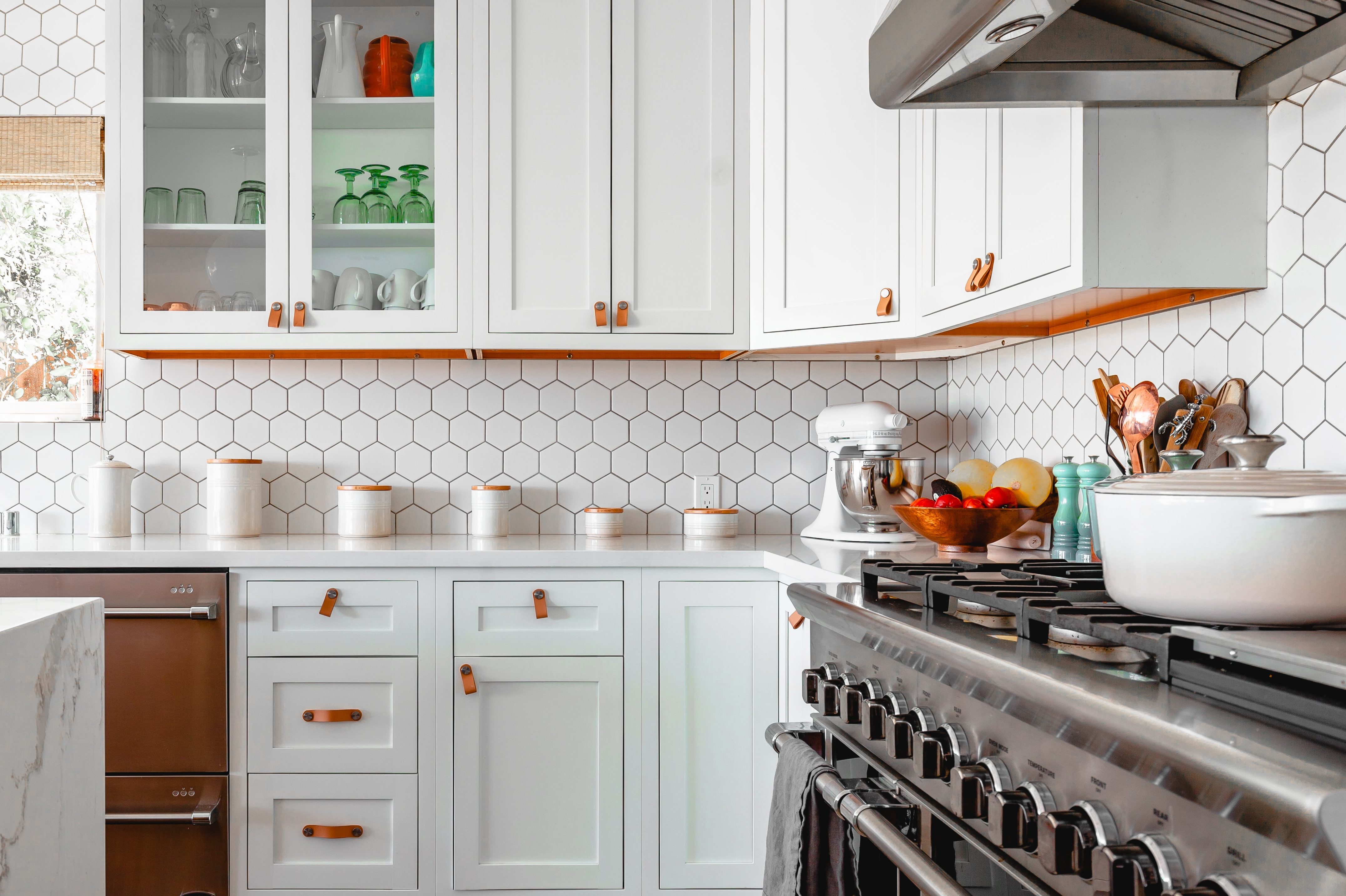 Brightly lit kitchen with white cupboards and white tiles and a silver gas stove featuring a French oven.