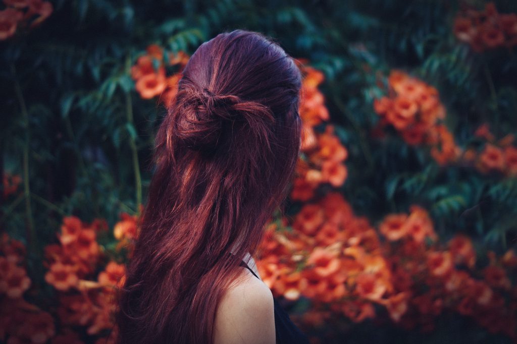 Girl with long red hair faces a wall of green leaves and orange-red flowers.