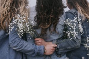 Three women embracing, viewed from behind. All three women have long hair and are wearing blue clothing, and are carrying small white flower bouquets.