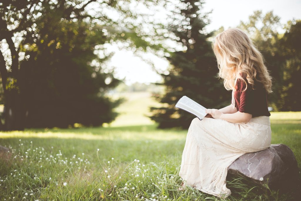 Girl with long blond hair wearing a dark red top and long white skirt sits relaxed on a rock holding a book, reading. A pasture with summer flowers in pine trees are in the background.