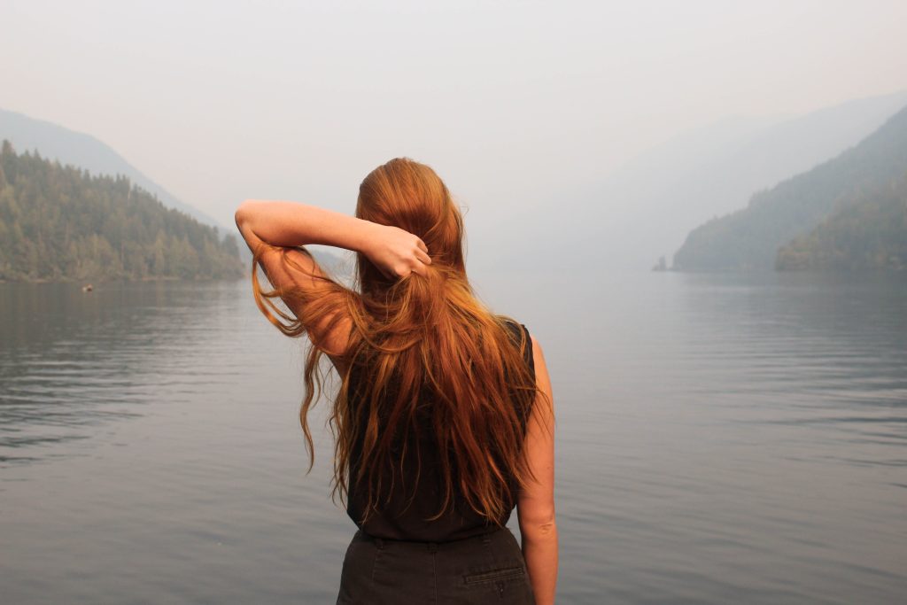 Girl with long red hair is facing a lake and mountains that are partially obscured by mist. She is holding some of her hair at the back of her head with her left hand.