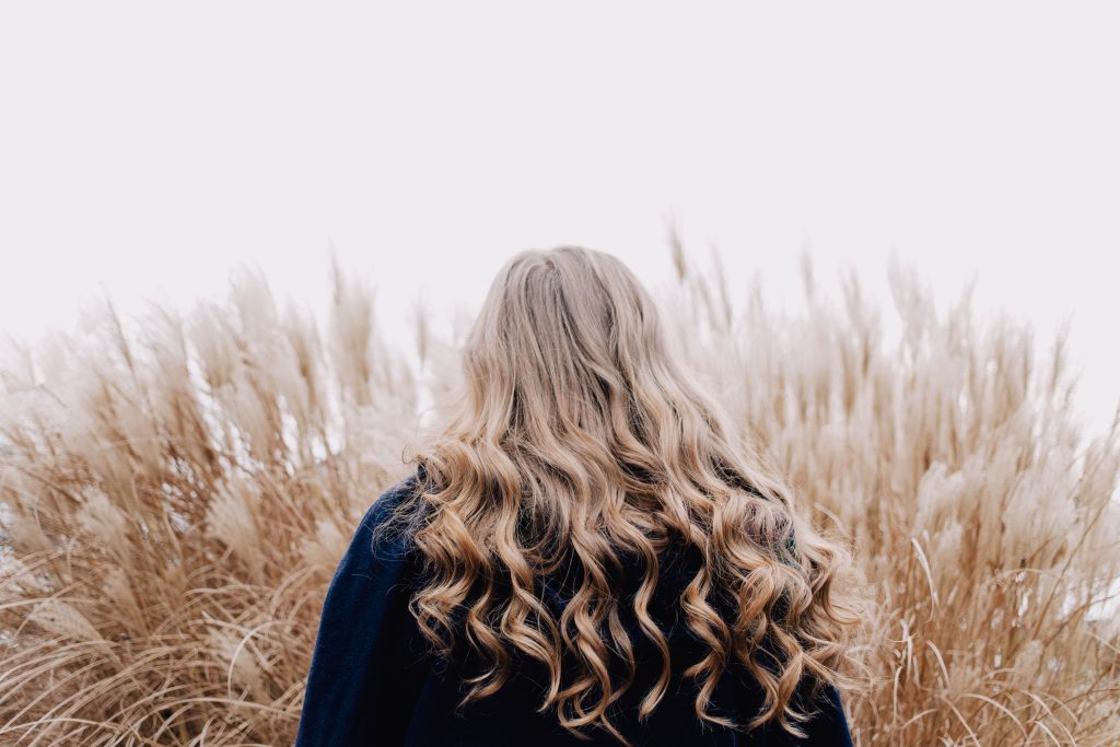 Woman with long blond curled hair is facing away. She is wearing a black top and is facing a stand of tall golden grasses.