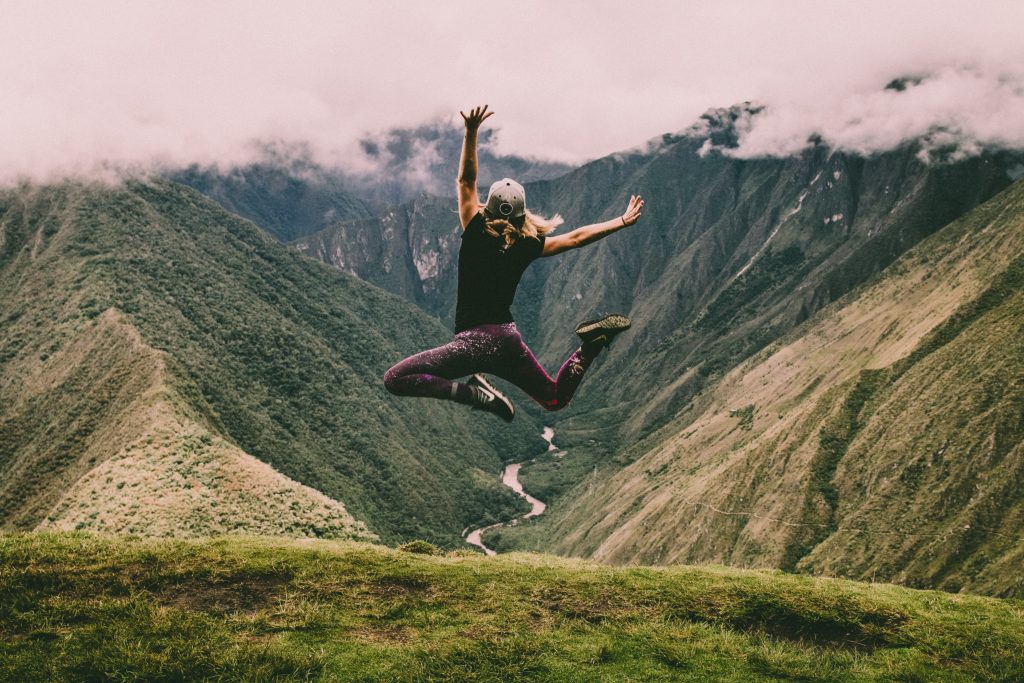 Girl in hiking clothes jumping up in the air facing green mountains. The mountain tops are hidden in clouds, a river is seen at the bottom.