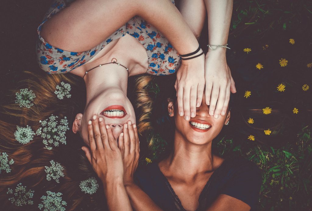 Two woman lying down on the grass with flowers in their hair. They are covering each other's eyes and smiling with red lipstick on.