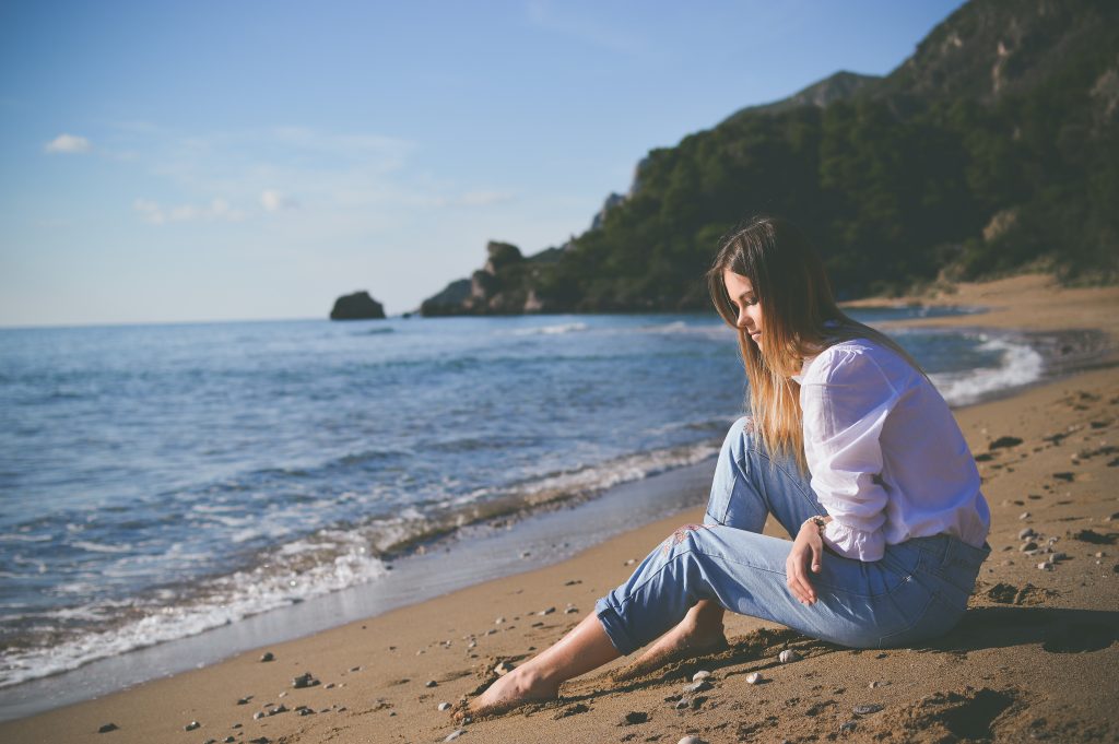 Girl with long brown hair wearing a white blouse and blue jeans is sitting barefoot on a sandy beach, facing the ocean.