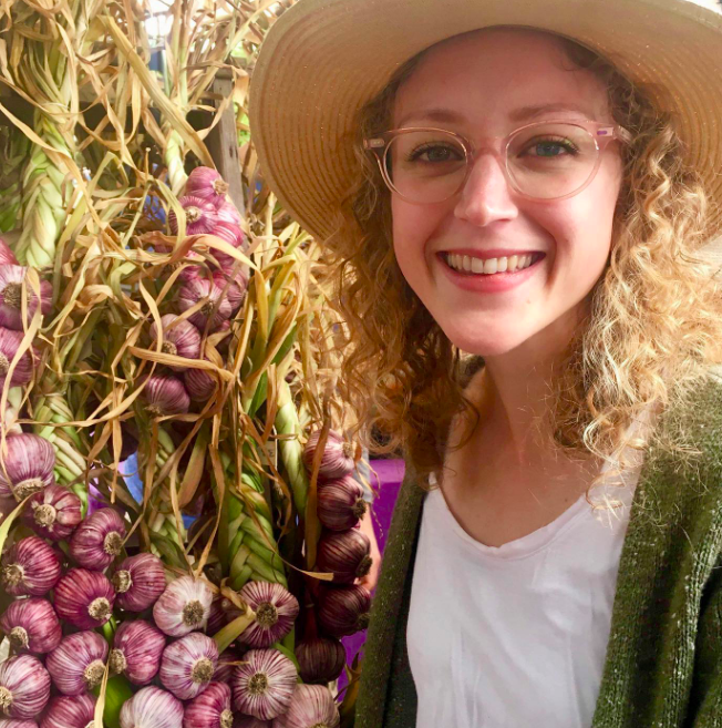 Girl with curly hair wearing a summer hat, standing next to braided purple garlic.