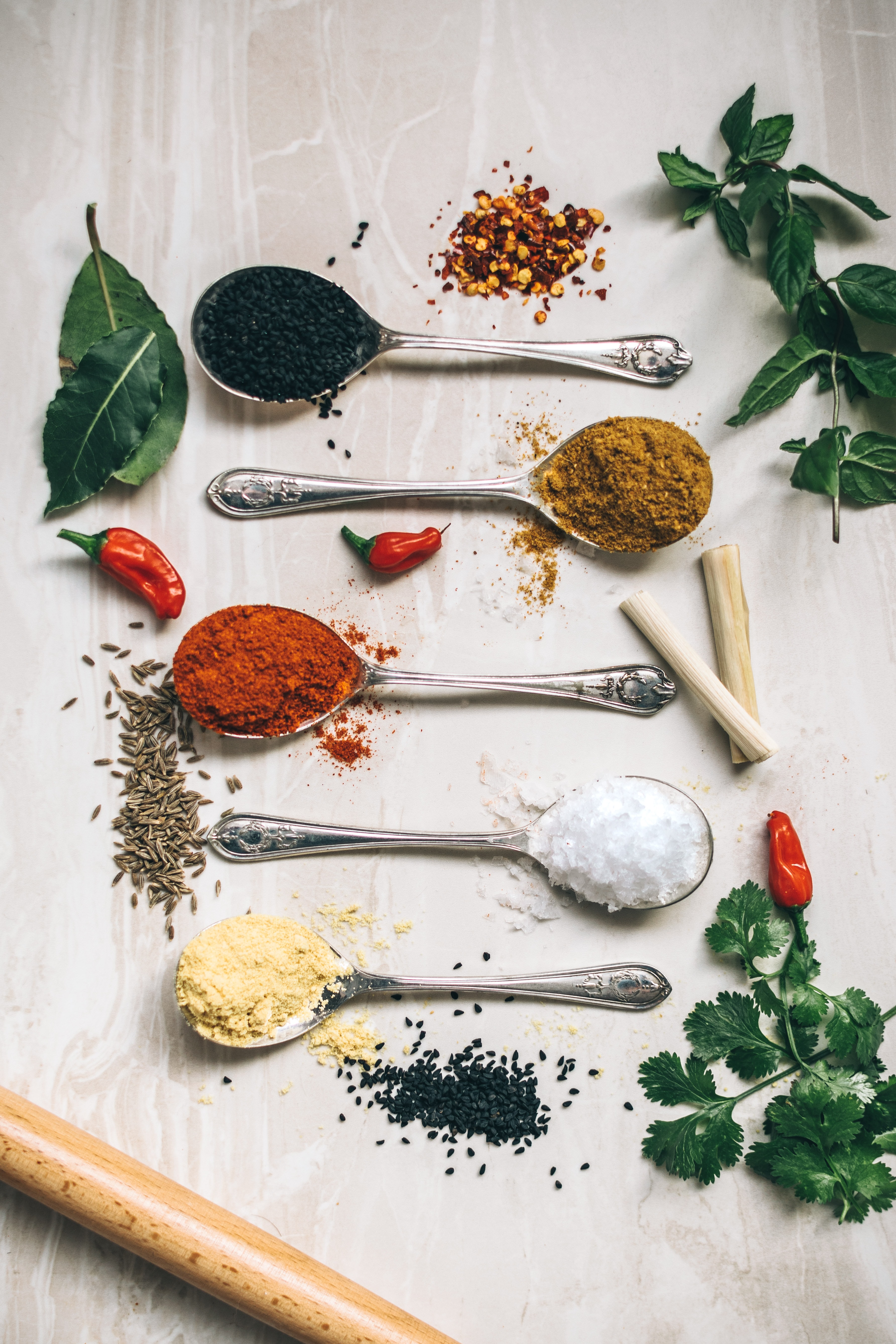 silver spoons aligned together with spices and herbs dispersed around them on a beige background.